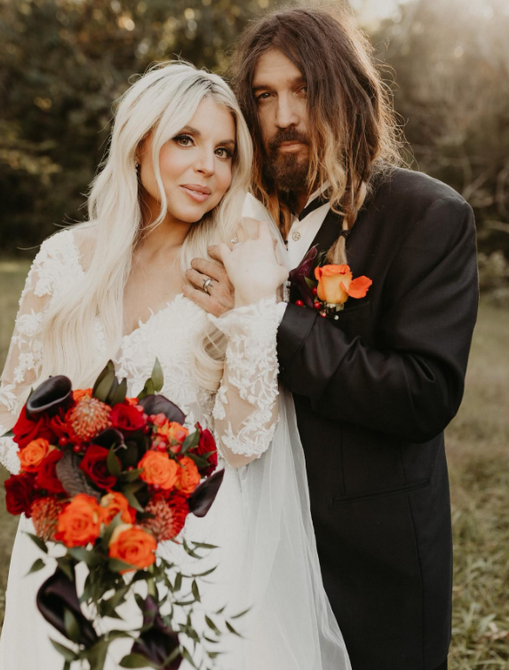 Billy Ray Cyrus and Firerose on their wedding day in October 2023. She is holding a red and orange bouquet and wearing a white dress. Cyris is wearing a black tux.